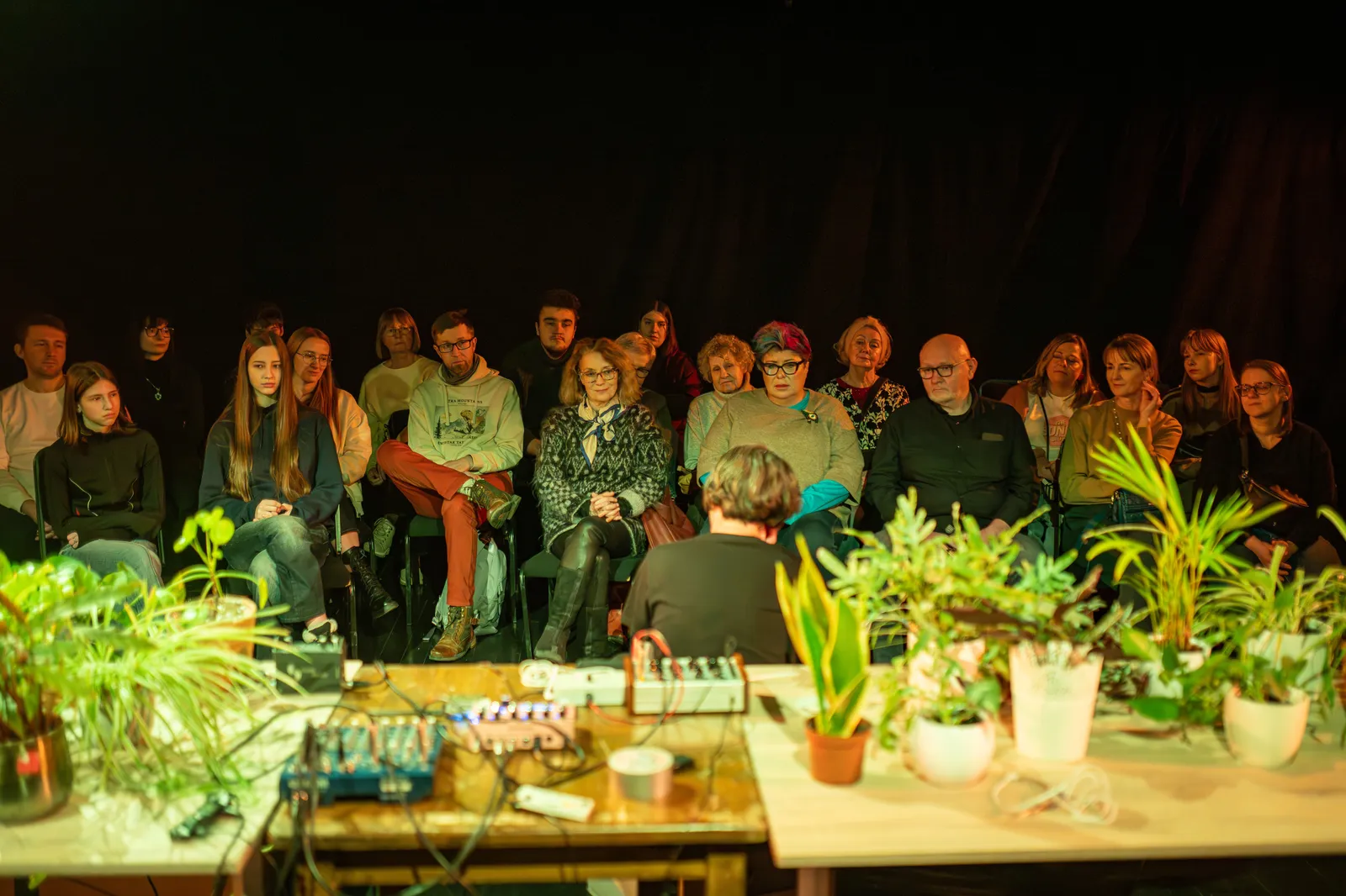 Osoba prowadząca wydarzenie stoi na scenie przed siedzącą publicznością; obok stół z roślinami doniczkowymi i sprzętem elektronicznym, oświetlenie teatralne.