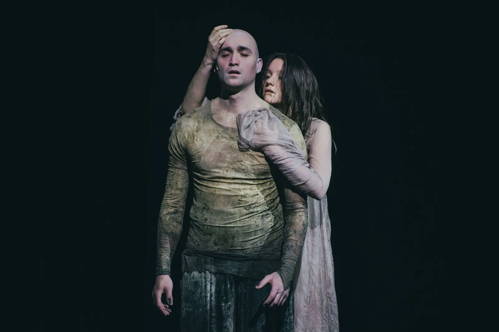 Na zdjęciu dwie osoby (Marek Puchowski, Maria Wróbel) na scenie teatralnej. Jedna stoi z zamkniętymi oczami, druga obejmuje ją od tyłu, dotykając głowy. Oświetlenie punktowe, ciemne tło.