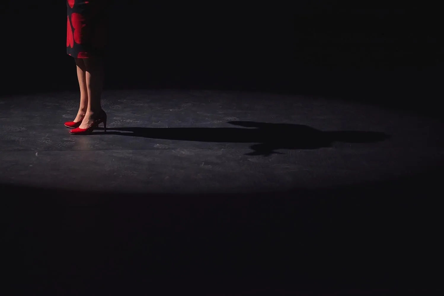 Fragment sceny teatralnej: widoczne tylko nogi kobiety w czarnej spódnicy i czerwonych szpilkach. Na podłodze wyraźny cień sylwetki, otoczony ciemnością i punktowym światłem.