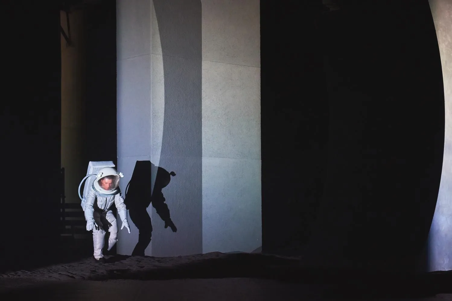 Postać w białym skafandrze astronauty porusza się po ciemnej scenie. Silne światło rzuca wyraźny cień na ścianę. Scenografia minimalistyczna, surowa, przypominająca przestrzeń kosmiczną.