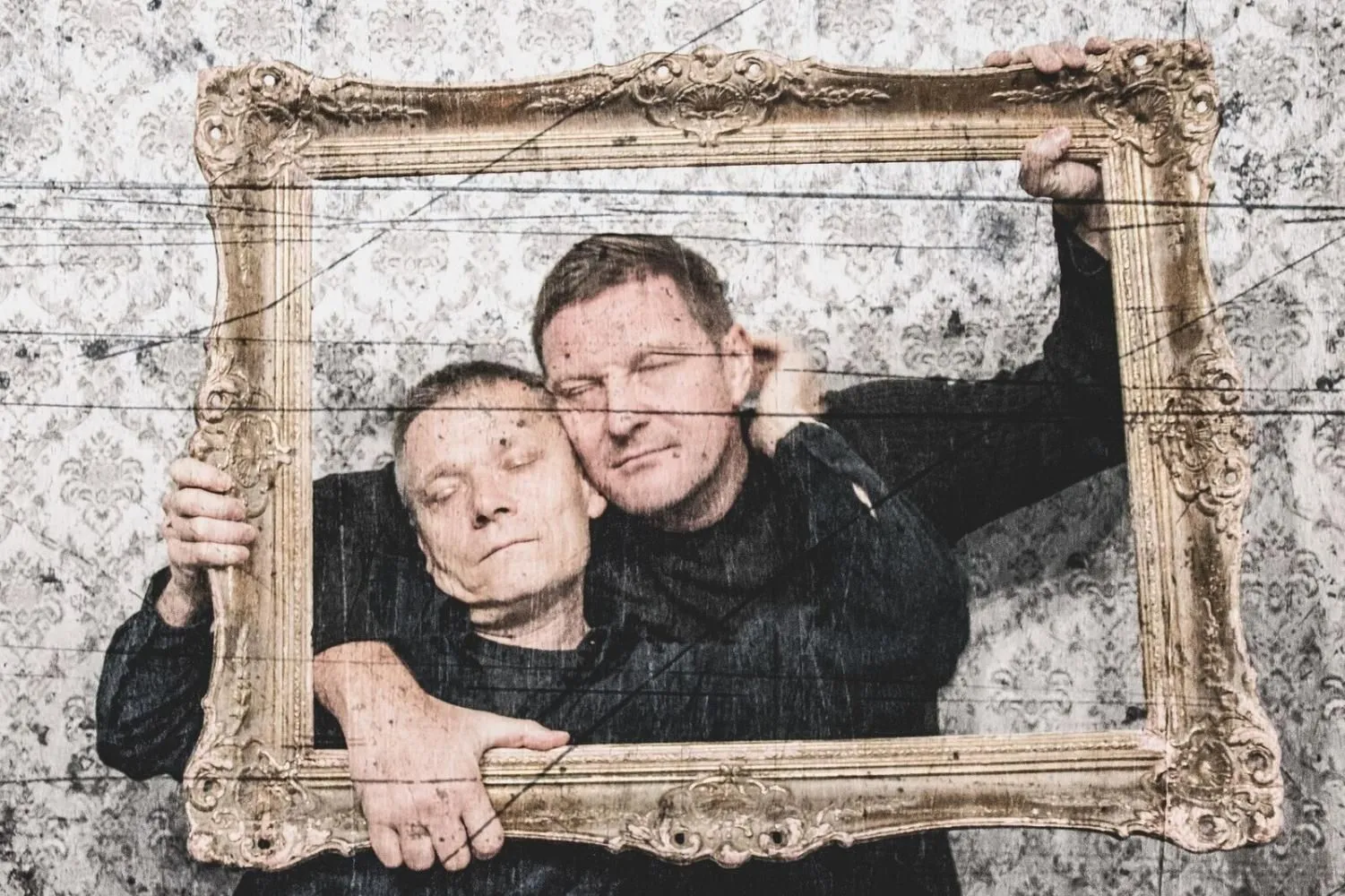 Dwaj mężczyźni ubrani na czarno obejmują się, trzymając pustą, ozdobną ramę obrazu. Ich twarze są blisko siebie, oczy zamknięte. Tło stanowi jasna, wzorzysta tapeta.