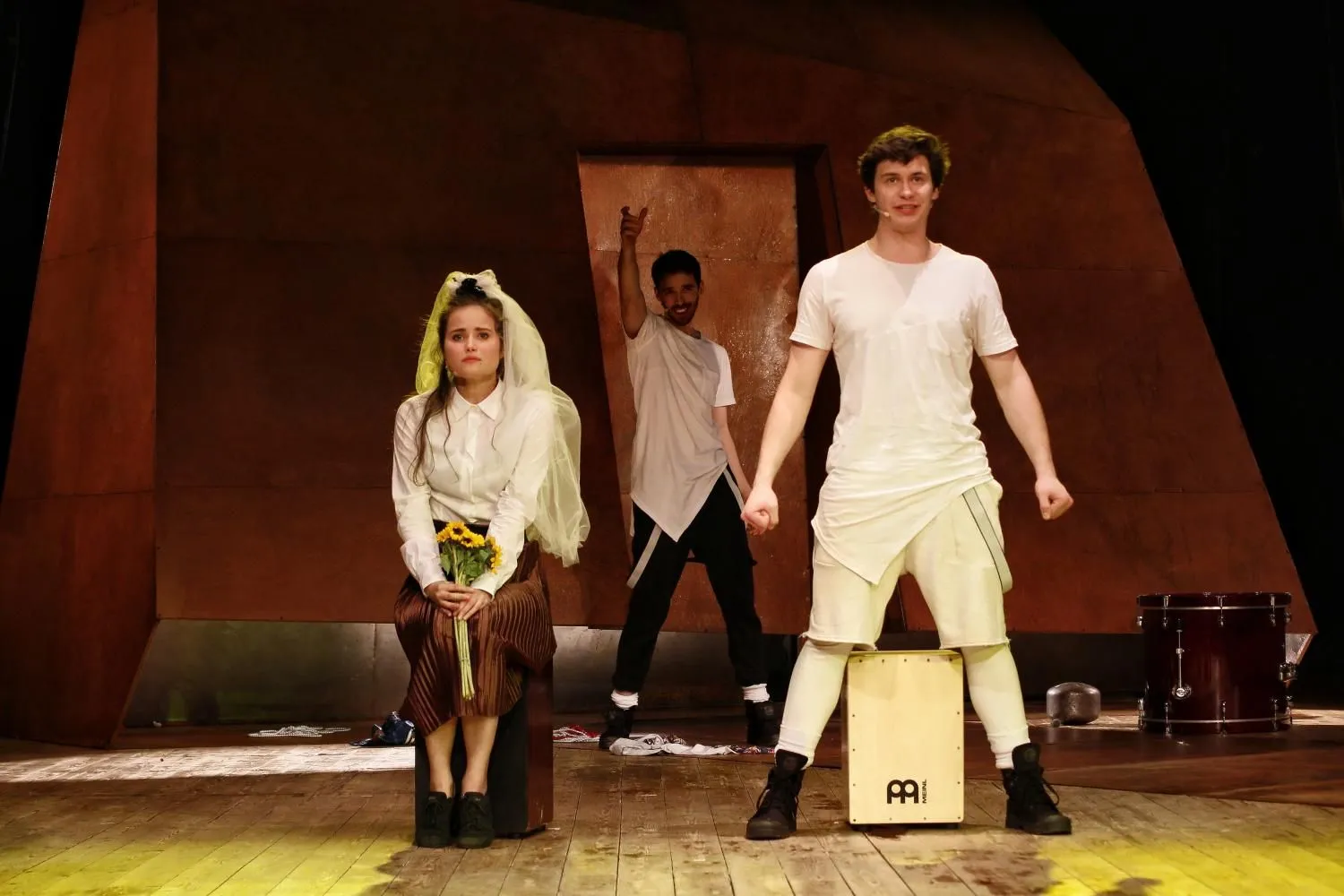 Scena teatralna: kobieta w białej koszuli i welonie siedzi z bukietem kwiatów, obok mężczyzna stoi z nogami postawionymi między skrzynką. W tle kolejny aktor w geście uniesionej ręki. Drewniana podłoga, brązowa scenografia.
