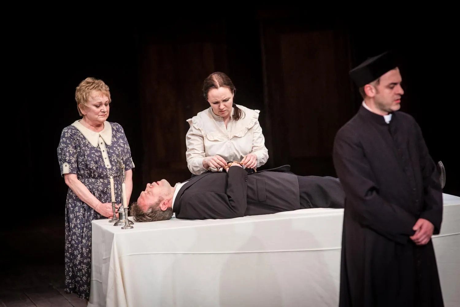 Scena teatralna: ciało mężczyzny leży na stole przykrytym białym obrusem. Dwie kobiety stoją przy nim, jedna poprawia ubranie zmarłego. Ksiądz odwrócony tyłem. Nastrój żałoby i skupienia.