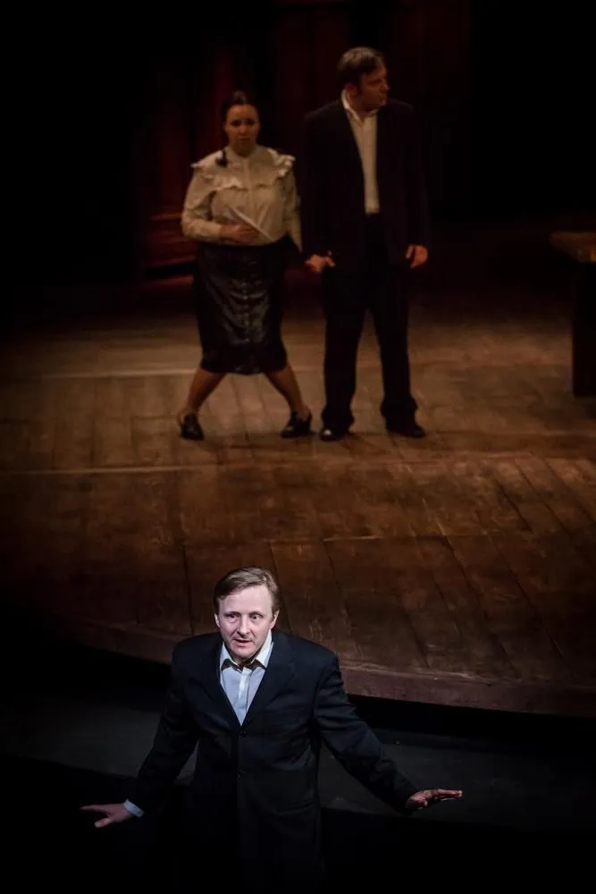 Scena teatralna: kobieta i mężczyzna stoją na drewnianej scenie, trzymając się za ręce. Na pierwszym planie inny mężczyzna zwraca się w stronę widowni. Minimalistyczna przestrzeń, skupienie na relacjach.