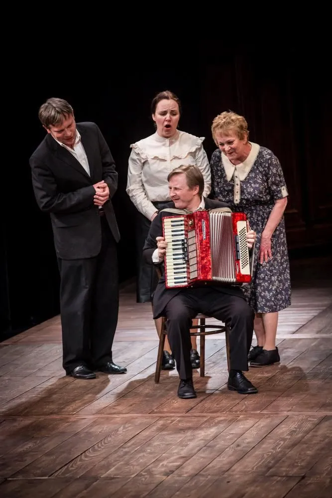 Scena teatralna: mężczyzna siedzi na krześle i gra na czerwonym akordeonie. Trzy osoby stoją wokół, słuchając lub śpiewając. Prosta scenografia, centralna rola muzyki.