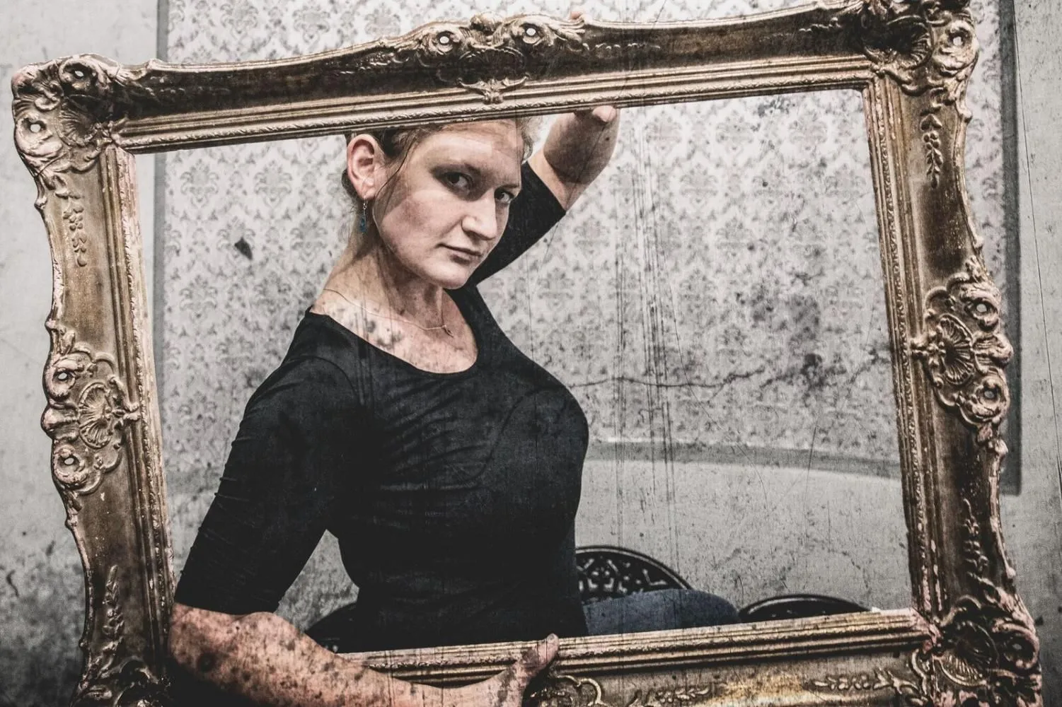 Kobieta w ciemnym stroju pozuje wewnątrz pustej, ozdobnej ramy obrazu. Jedną rękę opiera o górną krawędź ramy. Patrzy w stronę obiektywu. Tło to jasna, wzorzysta ściana.