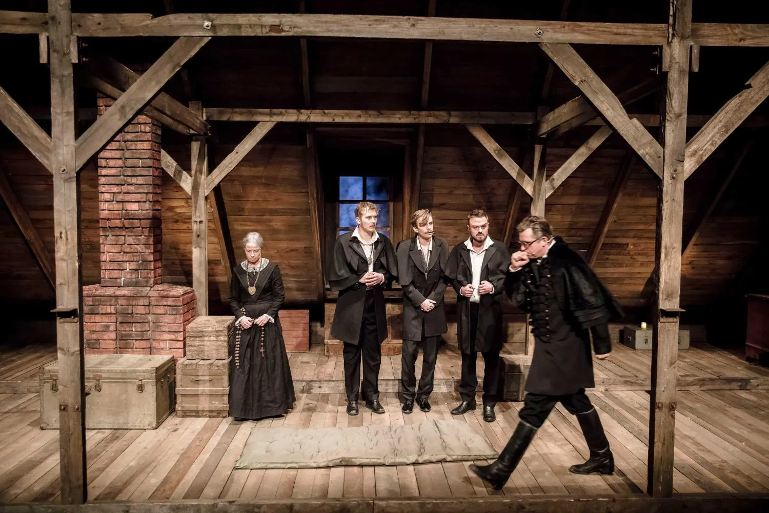 Kilka osób stoi w szeregu na środku sceny. Towiańska (Joanna Kreft-Baka) siedzi z boku na skrzyni. Towiański (Maciej Konopiński) w czarnym płaszczu przechodzi przed nimi.