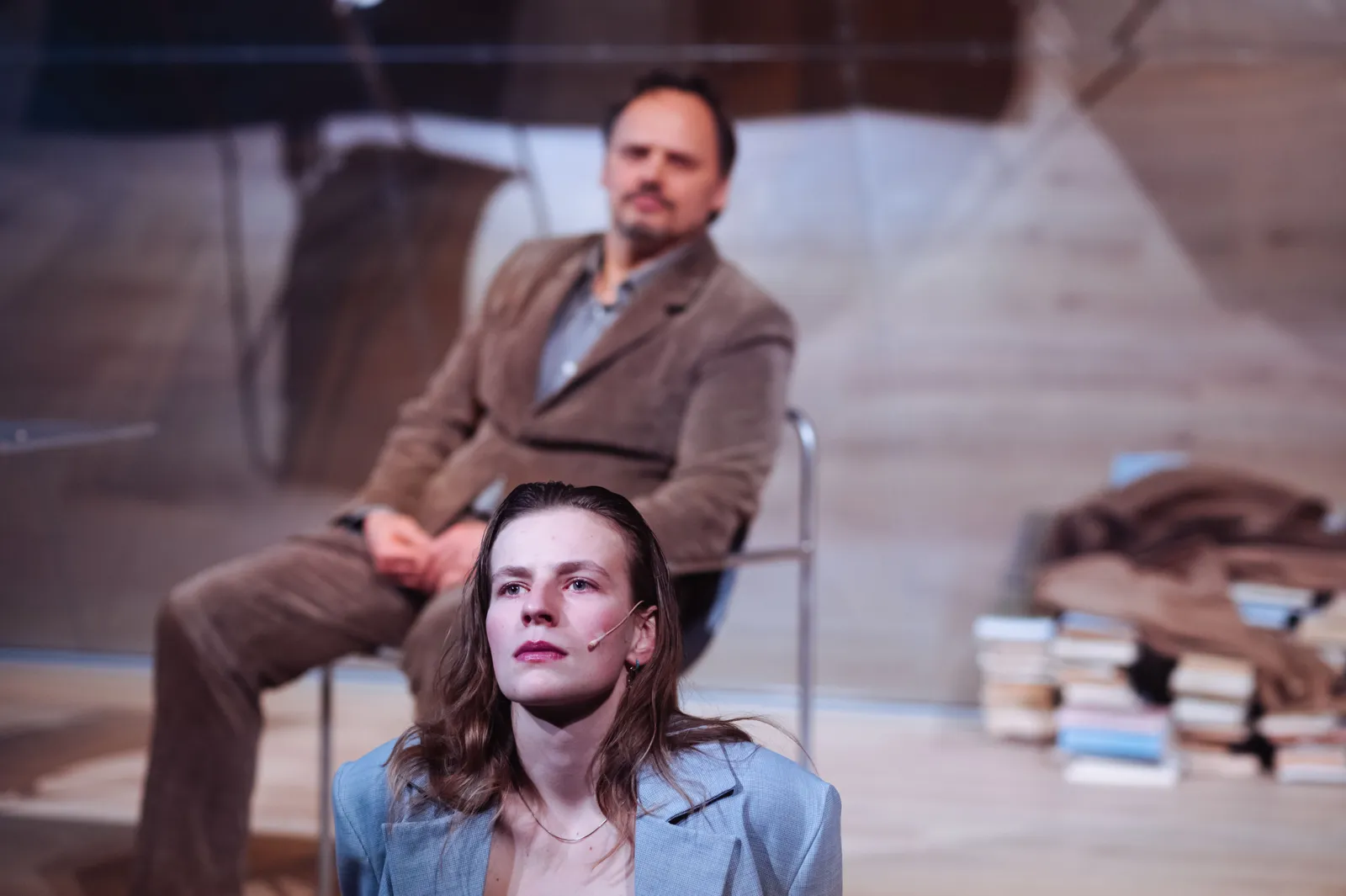 Aktorka siedzi na podłodze na pierwszym planie, przed nią pusta przestrzeń sceny; w tle mężczyzna siedzi nieruchomo przy ścianie, obok stos książek.