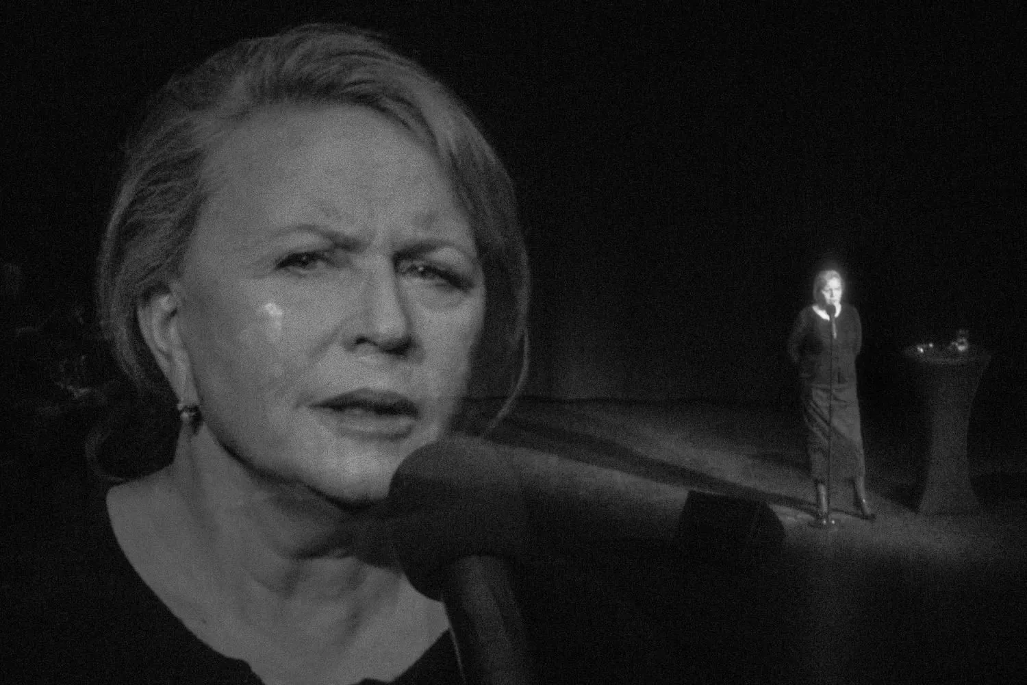 Duże zbliżenie twarzy kobiety mówiącej do mikrofonu; w tle widoczna jej sylwetka na scenie.