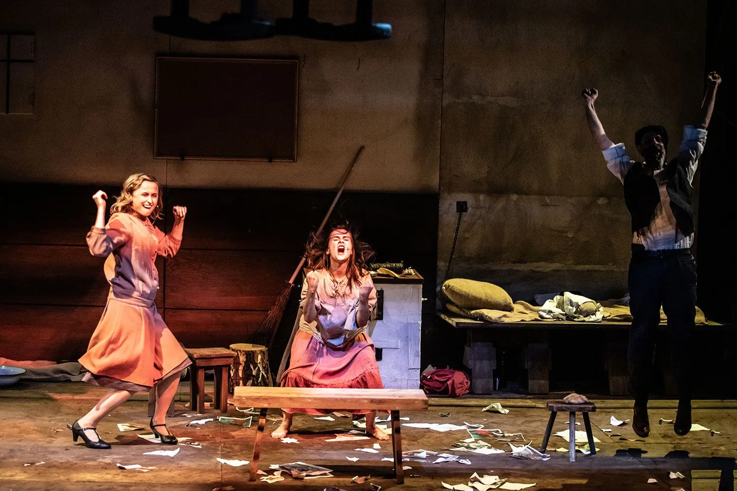 Na scenie dwie kobiety (od lewej: Justyna Bartoszewicz, Karolina Kowalska) w dynamicznych pozach; po prawej w cieniu skacze mężczyzna (Marek Tynda) z uniesionymi rękoma; wokół nich porozrzucane kartki i proste meble.