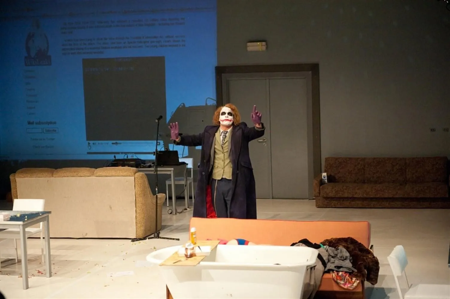 Postać w teatralnym makijażu stoi na środku sceny z uniesionymi rękami. W tle projekcja strony internetowej oraz elementy scenografii mieszkania.