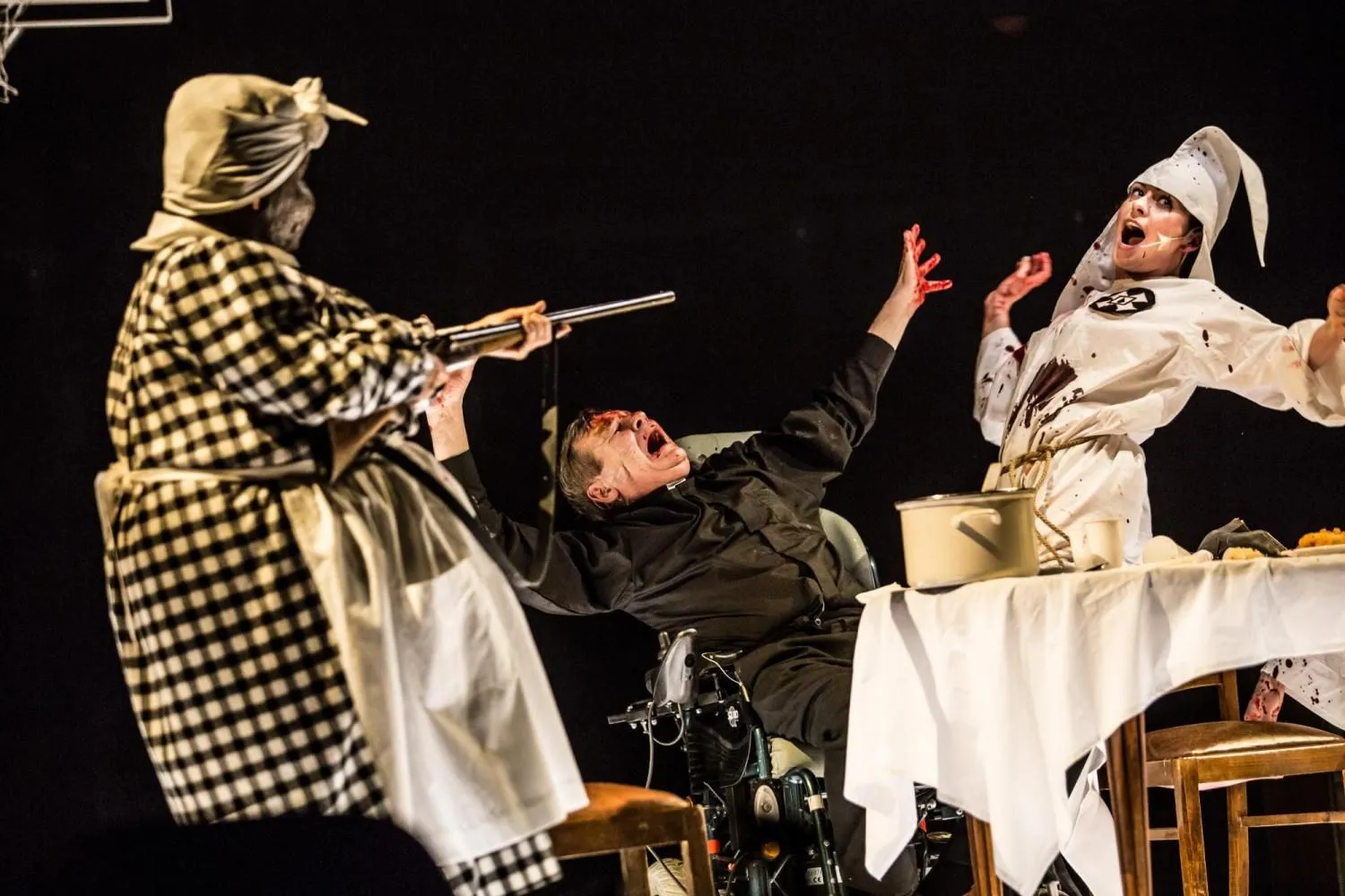 Scena gwałtownego gestu: jedna postać mierzy długą bronią w stronę siedzącego mężczyzny, inna reaguje krzykiem; teatralne światło podkreśla napięcie sytuacji.