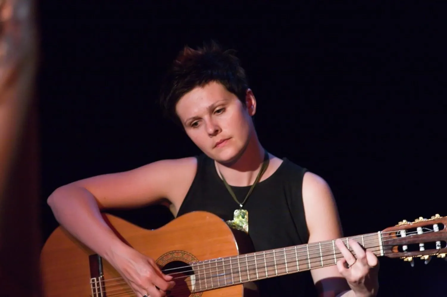 Dziewczyna z krótkimi włosami gra na gitarze. Ma czarną koszulkę bez rękawów i duży naszyjnik. Patrzy z natchnieniem w dół.