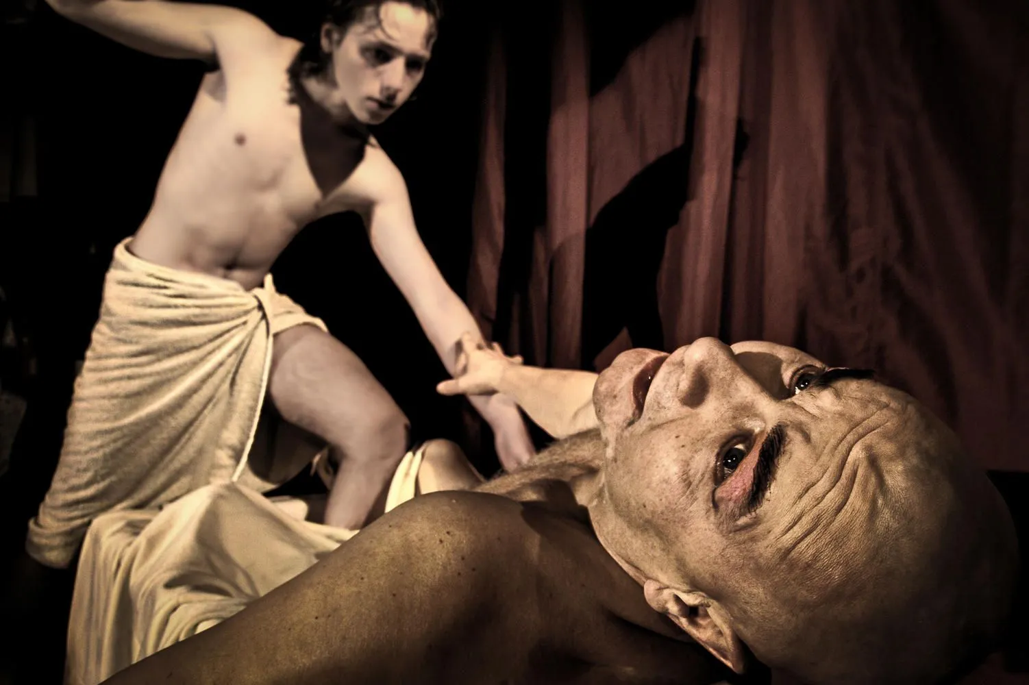 Scena teatralna przedstawia dwóch półnagich mężczyzn. Jeden leży na plecach, patrząc w górę z napiętym wyrazem twarzy, drugi pochyla się nad nim i chwyta go za rękę. Postacie są owinięte jasną tkaniną, tło ciemne, światło skupione na ciałach.