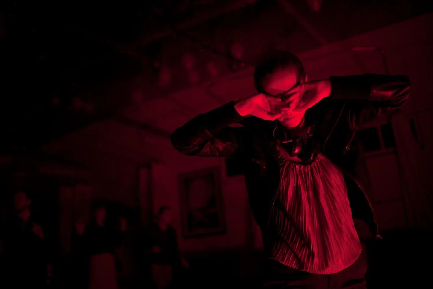 Performer stoi na scenie w czerwonym świetle, z rękami przy twarzy, jakby zasłaniał oczy. Ma na sobie ciemną kurtkę i jasną koszulę. Tło jest mroczne i rozmyte, co podkreśla teatralny, niepokojący nastrój sceny.