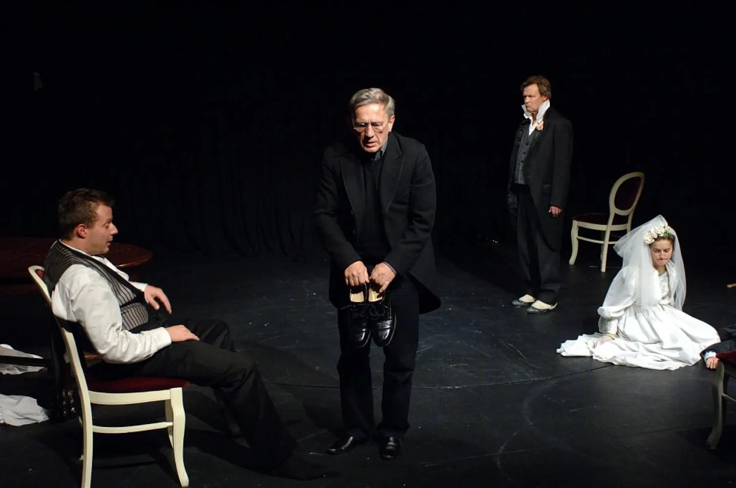 Scena teatralna w ciemnej przestrzeni. Na pierwszym planie mężczyzna w czarnym garniturze trzyma w dłoniach parę czarnych butów. Po bokach siedzą i stoją inni aktorzy, w tle kobieta w białej sukni ślubnej siedzi na podłodze.