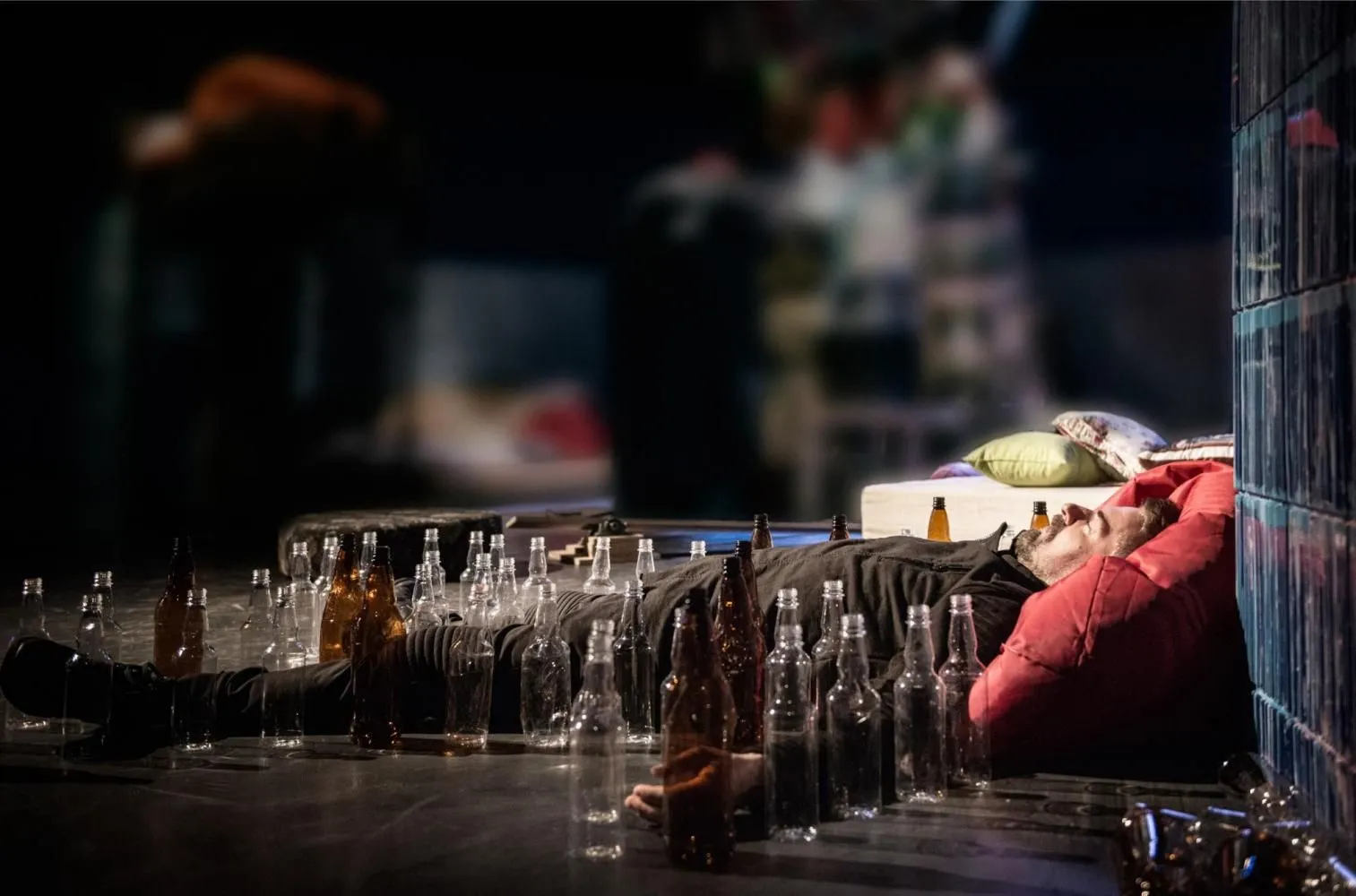 Mężczyzna leży na scenie oparty o ścianę, otoczony pustymi i pełnymi butelkami; obok poduszki i elementy scenografii sugerujące zagracone wnętrze.