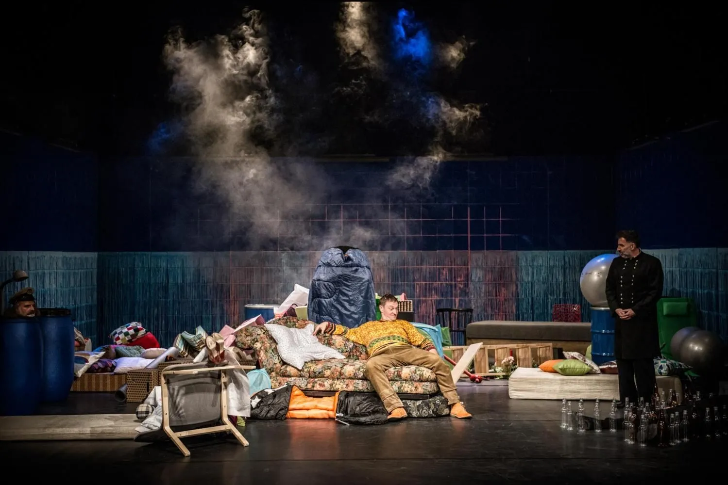 Szeroki plan sceny: mężczyzna siedzi na kanapie pośród porozrzucanych przedmiotów, inny stoi z boku; nad sceną unoszą się kłęby dymu w teatralnym świetle.