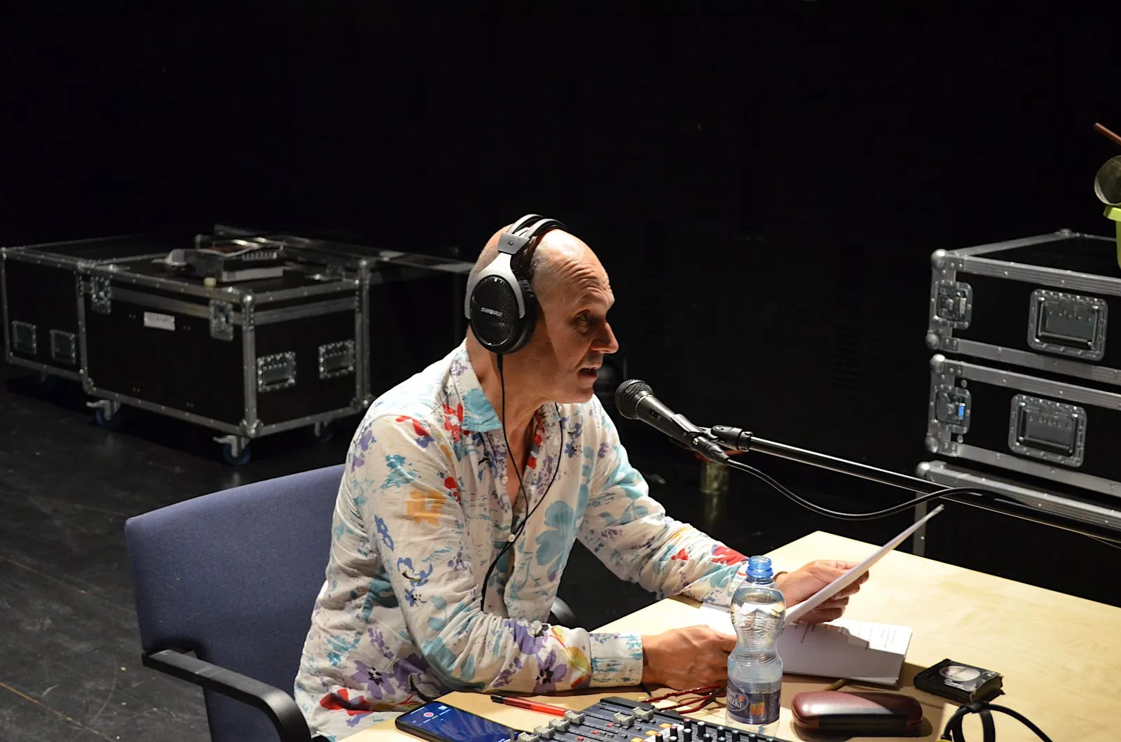 Lektor audiodeskrypcji w trakcie odczytu. Jest łysy, ma kwiecistą koszulę. Na uszach słuchawki, przed nim mikrofon, czyta z kartki.