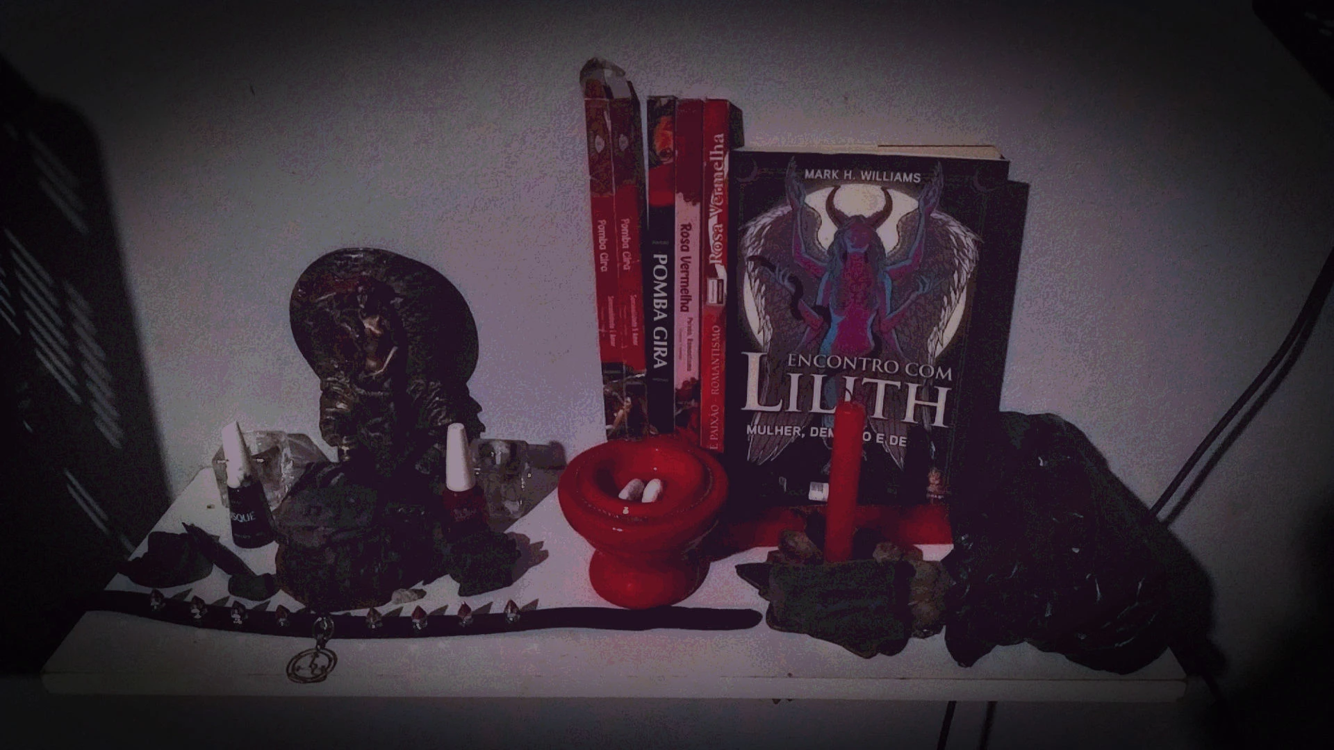 Heavily edited photography centered on a wall shelf containing plenty of ritualistic objects, from right to left: - A small, black bag (contains fresh red and black candles, not visible on picture). - Book "Encontro com Lilith: Mulher, Demônio e Deusa" (Meeting with Lilith: Woman, Demoness and Goddess) from Mark H Williams (a writer and bishop). The book is standing against the wall. - Before the book, there's a 7-pointed star, Lotus flower shaped, brass candle holder, with a red candle stick on it. The candle is not lit (yet). Two small pieces of charcoal are surrounding the candle. - A few incense stick boxes: two "Rosa Vermelha" (Red rose) from different brands (Darshan and Parman), a "Pomba Gira" (feminine spirits from Umbanda/Quimbanda) dedicated incense, and an empty incense stick box from a different brand which also reads "Pomba Gira: Sensualidade e Amor" (Sensuality and Love). All the incense stick boxes are vertically positioned against the wall. - Before the incense stick boxes, another candle-incense holder, this one being a red, circular porcelain/ceramic. Inside the candle holder, there are two small chalk pieces. - A small, brass statue of Aphrodite/Persephone/Inanna, with the figure standing naked (but the nudity is too small, too shaded by the low light conditions, and too bleached by the photo filters, so it's not visible) on top of a sea shell (doubleas as a Lotus flower) against a circular background (doubles both as a Full Moon and as a halo) while the left arm covers the genitalia and the right arm brushes her own hair. Also as part of the statue, a lengthy serpent is coiled all the way from her legs to her right arm then finally hanging at the chest level while facing us with a hissing mouth. The figure's hair is painted red, and the body has a slight shade of pale red as well. By her feet, more pieces of charcoal, including a razor-blade-shaped charcoal carved with the Hebrew letters for "Lilith". The brass statue is flanked by two cubic crystal candle holders, and also by two nail polishers: a black "Risqué" to her right and a red "Colorama" to her left. - Further before the statue, a spiked choker with a silver pendant containing the Seal of Lilith. The tips of the spikes are all painted red.
