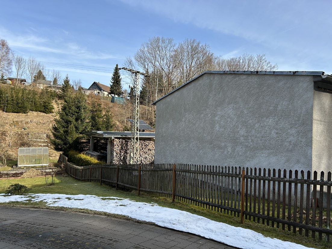 Gartenbereich mit Holzzaun, Schneeresten auf Rasen, Steinhaufen unter Dachüberstand und Blick auf bewaldeten Hang mit Wohnhäusern in Olbernhau, Rungstockstraße 120