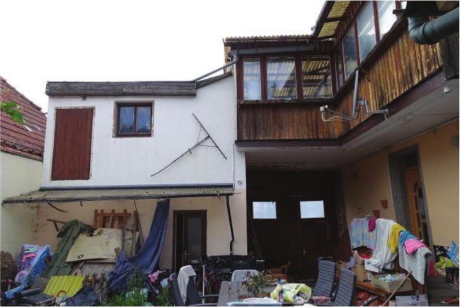 Rückansicht eines Einfamilienhauses mit Balkon, Fenstern und unordentlichem Hofbereich mit Gegenständen und Wäsche