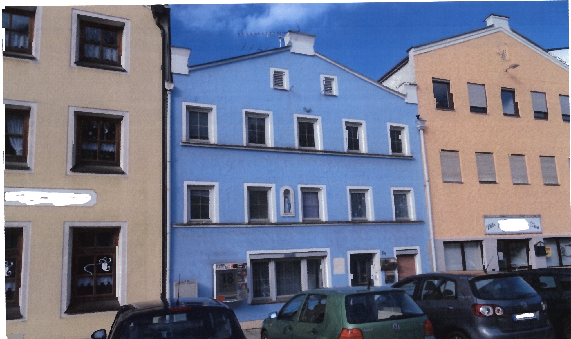 Fassade eines Wohn- und Geschäftshauses in Tann, Marktplatz 34, mit blauem Mittelteil und beigen Seitenflügeln, mehrere Fenster, geparkte Autos vorne