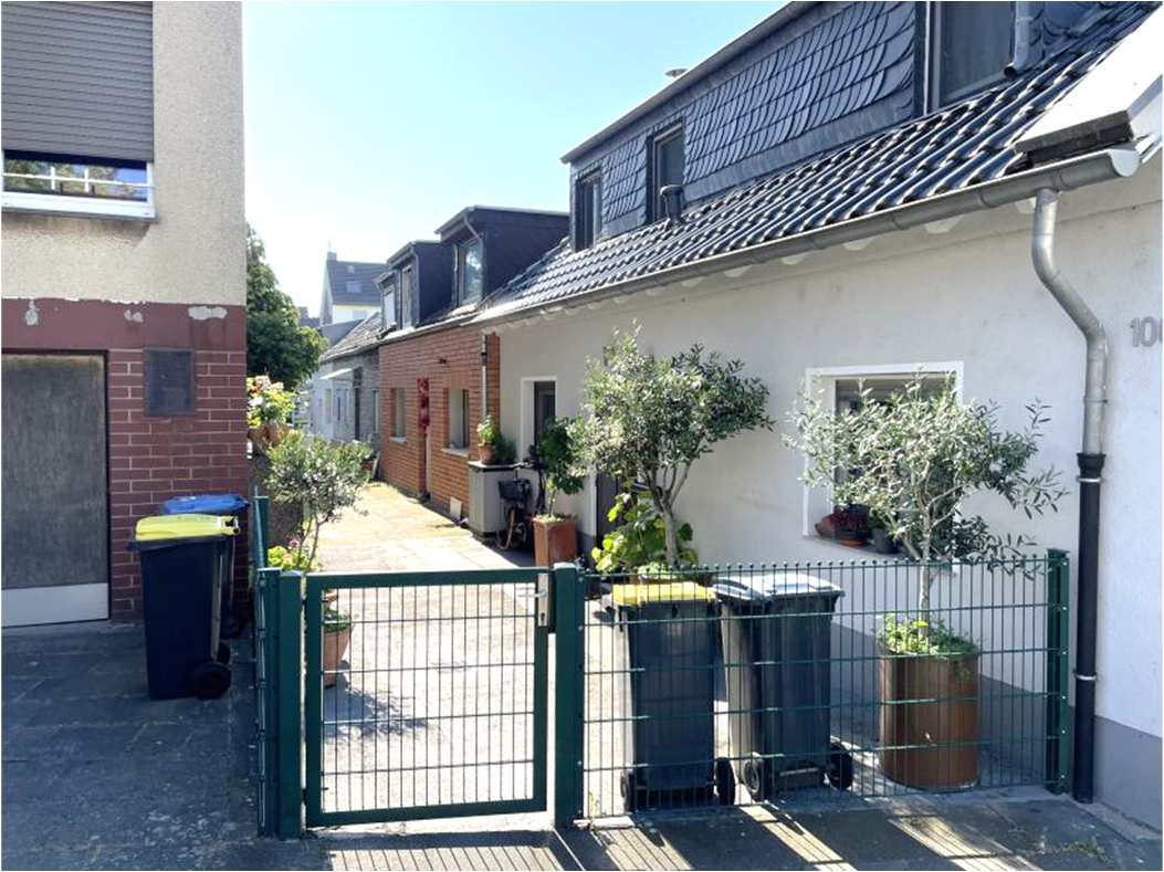 Hinterhofansicht eines Einfamilienhauses mit grünem Gittertor, Mülltonnen und Pflanzen in Kübeln, umgeben von Wohnhäusern in Köln-Niehl
