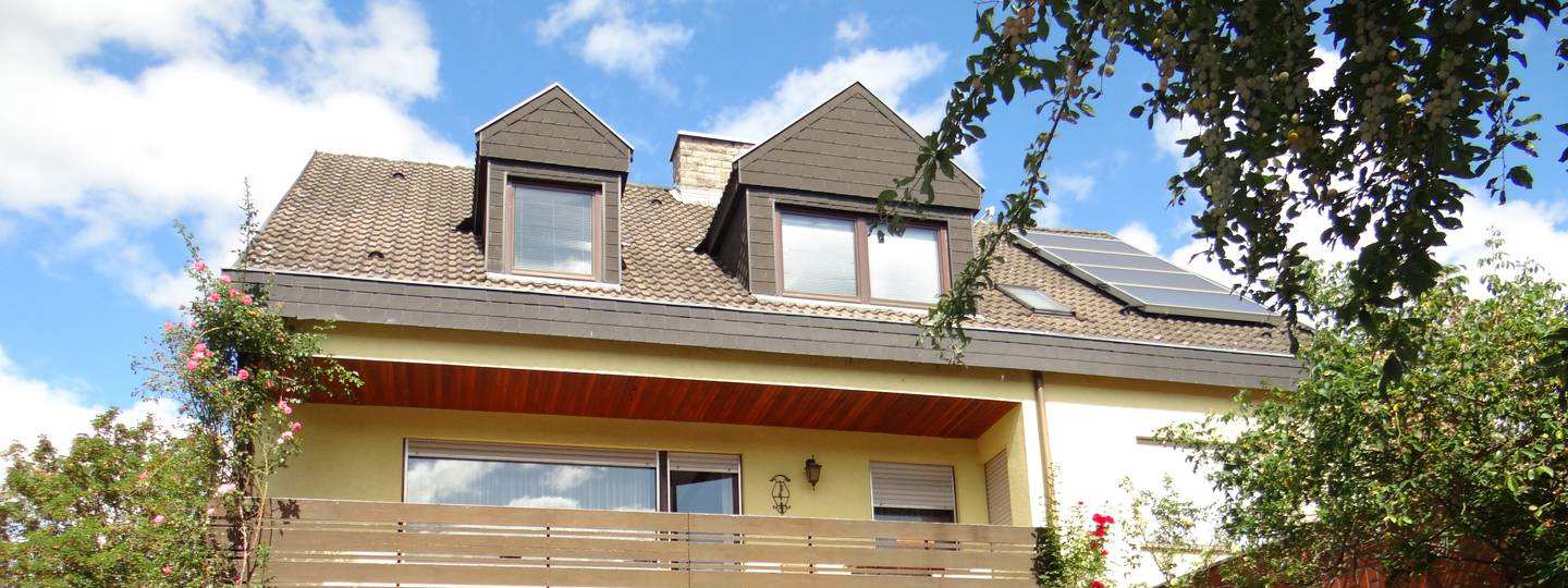 Einfamilienhaus mit gelber Fassade, Dachgauben und Solaranlage, umgeben von Bäumen und Sträuchern, Ansicht von vorne