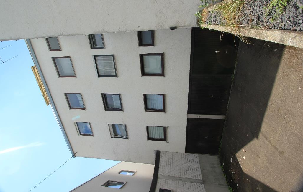 Eingangsbereich eines Gebäudes in Neu-Ulm, Arnulfstr. 12, mit mehreren Fenstern und einer dunklen Tür, gepflastertem Weg und angrenzender Mauer