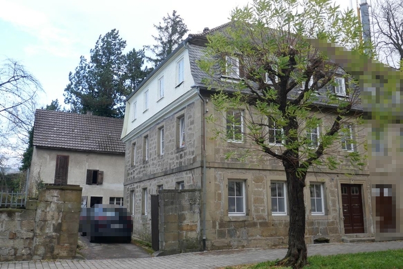 Fassade eines mehrstöckigen Steinhauses in Öhringen, Karlsvorstadt 35, mit grauem Satteldach, Fenstern und Eingangstür; vorne Baum und gepflasterter Hof