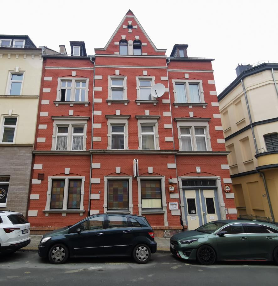 Rotes Backsteingebäude mit weißen Verzierungen, mehreren Fenstern und Eingangstür, Sedanstraße 5, Hof, 95028. Davor geparkte Autos