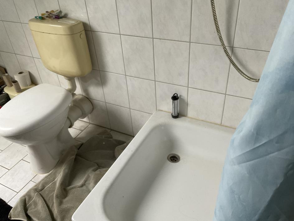 Badezimmer mit weißer Fliesenwand, WC mit cremefarbenem Spülkasten, Badewanne mit Duschschlauch und blauer Duschvorhang. Auf dem Boden liegt ein grauer Handtuch