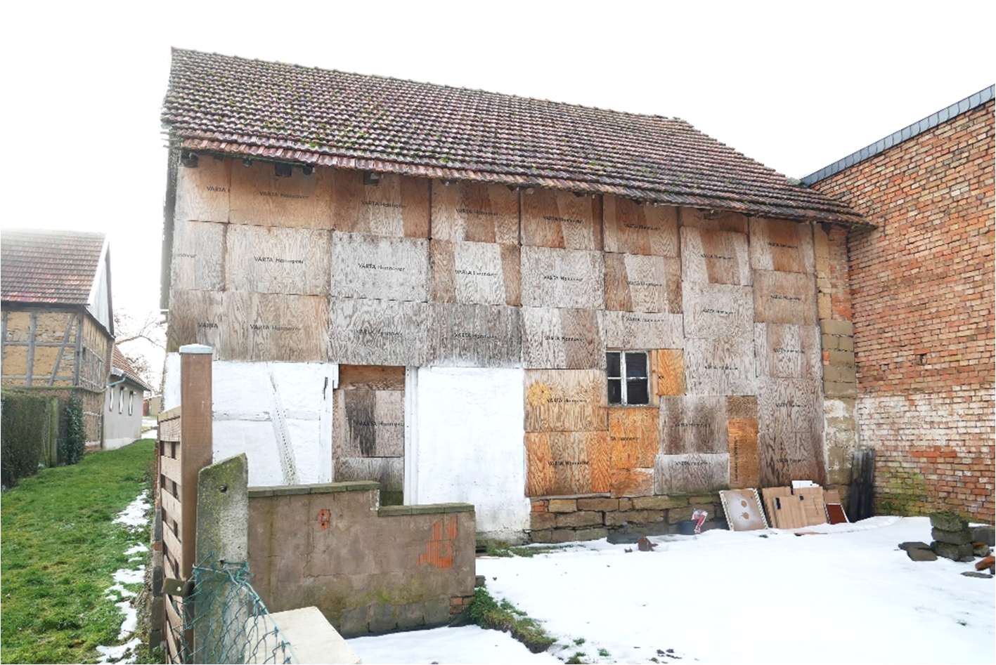 Seitenansicht eines Gebäudes in Freienbessingen mit Holzverkleidung, teilweise verputzt, Dach mit Ziegeln, Schnee am Boden, angrenzende Mauer und Grünfläche
