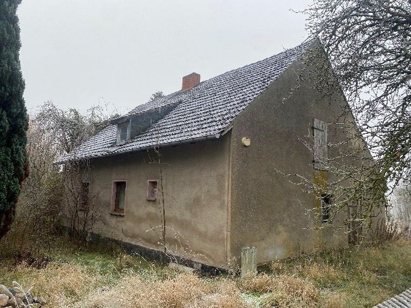 Einfamilienhaus in Zechin, Chausseestraße 9, mit grauer Fassade, Schieferdach und umgebendem Grün. Gebäude wirkt verlassen, Fenster teilweise vermauert