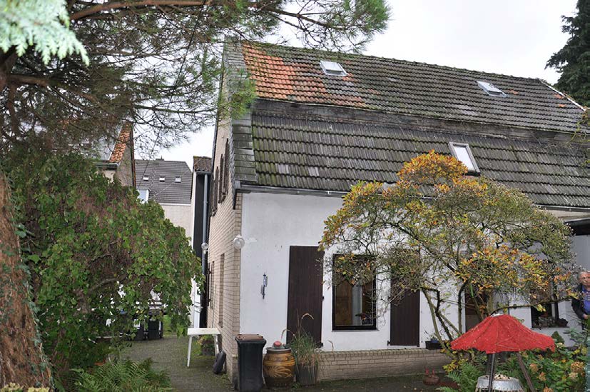 Seitenansicht eines Einfamilienhauses mit Ziegeldach, weißer Fassade, Fenstern und angrenzendem Gartenbereich mit Bäumen und Pflanzen