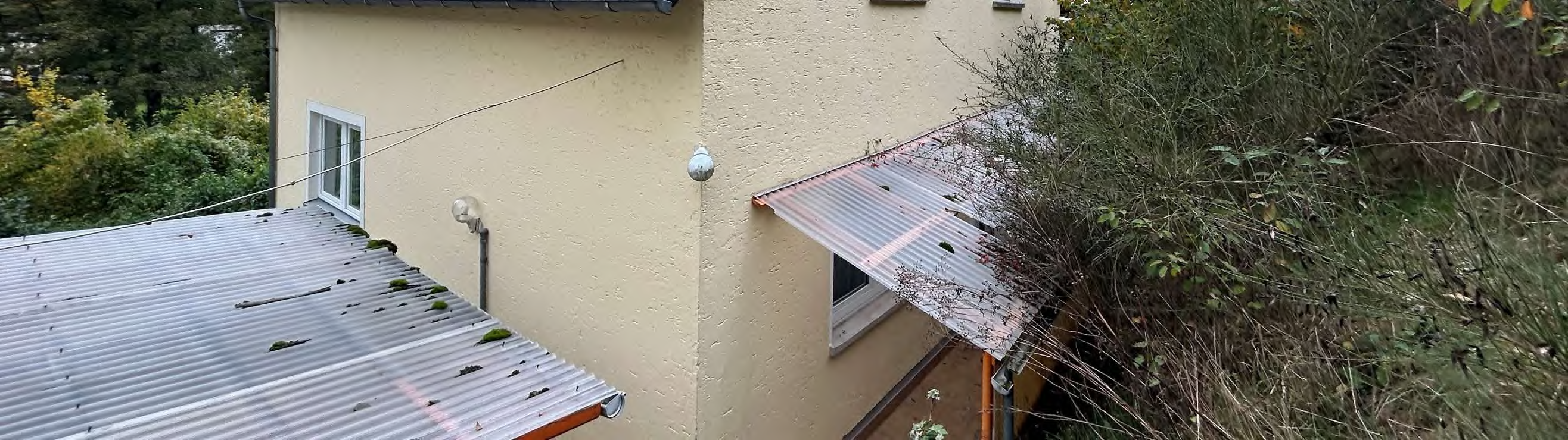 Seitenansicht eines Einfamilienhauses mit Wellblechdach, gelber Fassade und Fenster, teilweise von Sträuchern umgeben