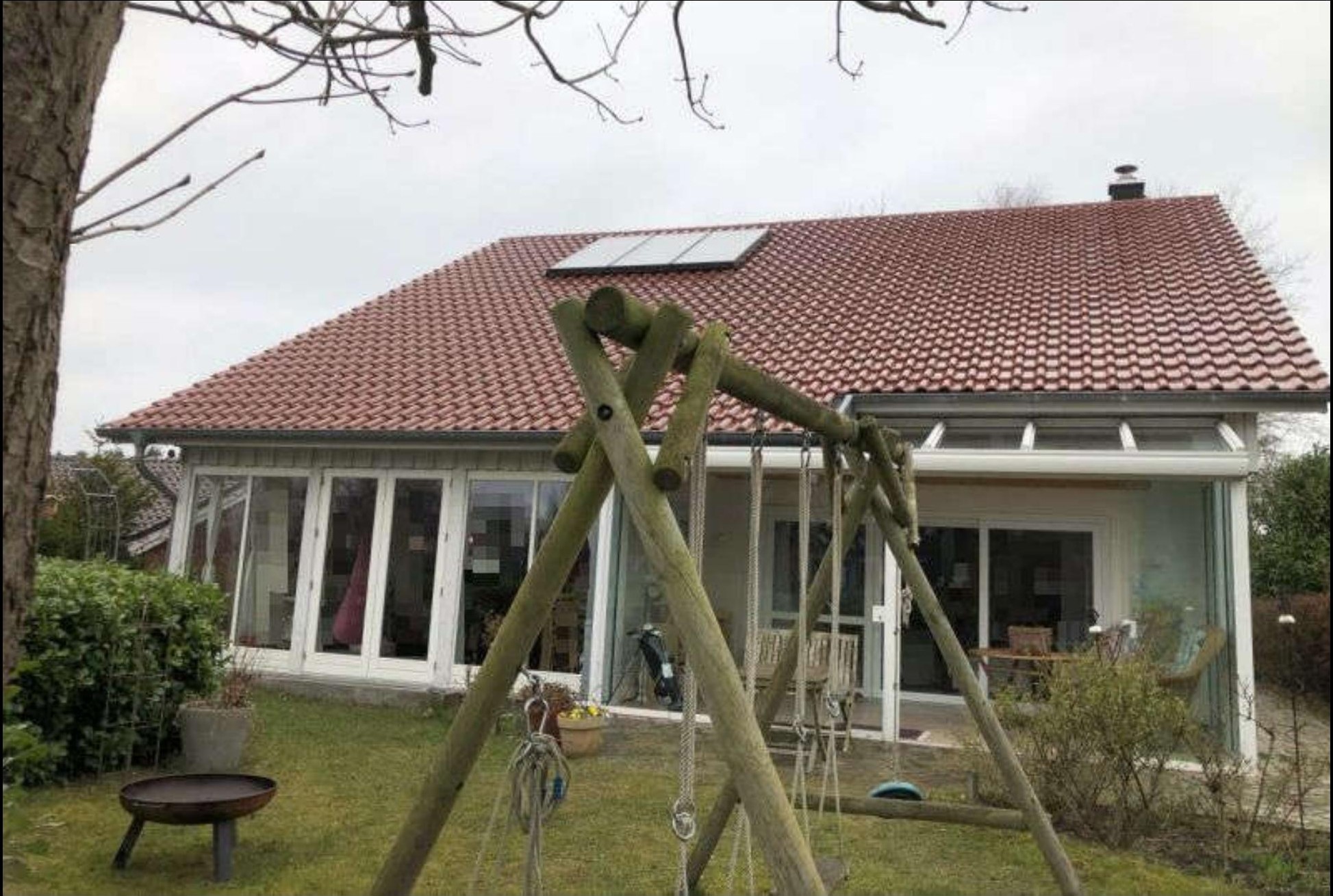 Gartenbereich mit Holzschaukel, Grasfläche, Sträuchern und Feuerschale vor einem Einfamilienhaus mit großflächigen Fenstern und Satteldach