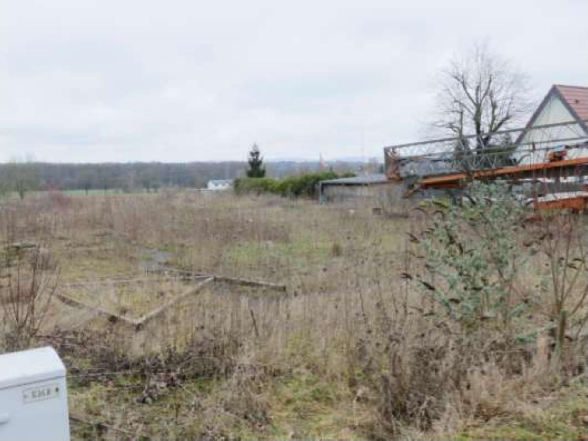 Überwuchertes Grundstück mit trockenem Gras, Holzrahmen und Blick auf ein Gebäude im Hintergrund in Löhne, Ellerbuscher Straße 128
