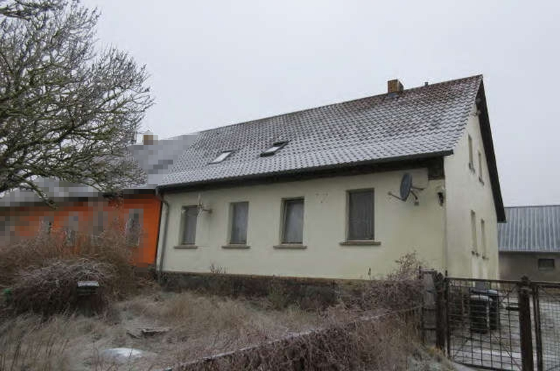 Einfamilienhaus in Oberuckersee, Dorfstr. 10, mit hellgelber Fassade, grauem Wellblechdach und mehreren Fenstern; umgeben von trockenem Gras und einem Metallzaun