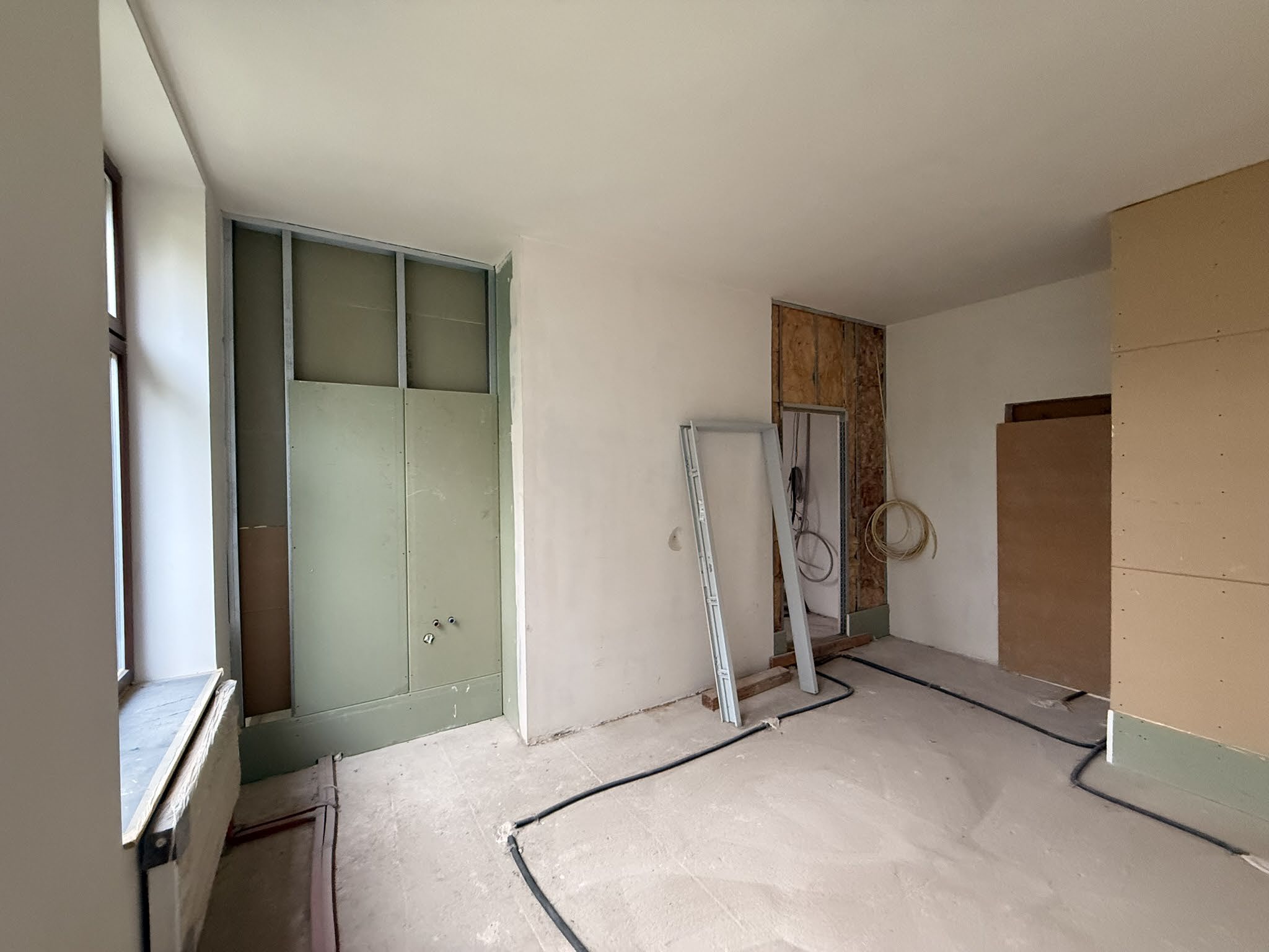 Unfertiger Innenraum einer Gewerbeeinheit mit sichtbaren Rohbauarbeiten, Metallrahmen, Holzverkleidung und Kabel auf dem Boden. Fenster links, offene Türöffnung im Hintergrund