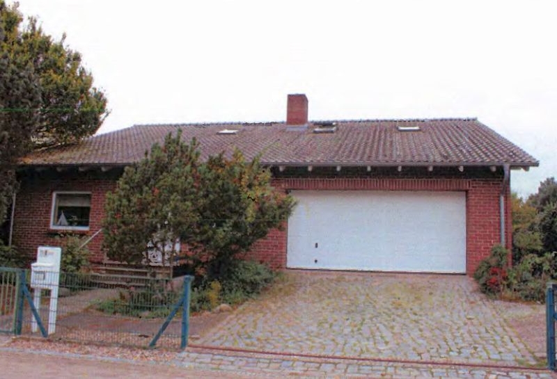 Einfamilienhaus mit rotem Ziegelmauerwerk, Satteldach mit Dachfenstern, weißer Garagentür und Kiesvorplatz. Links Briefkasten, umgeben von Bäumen und Sträuchern
