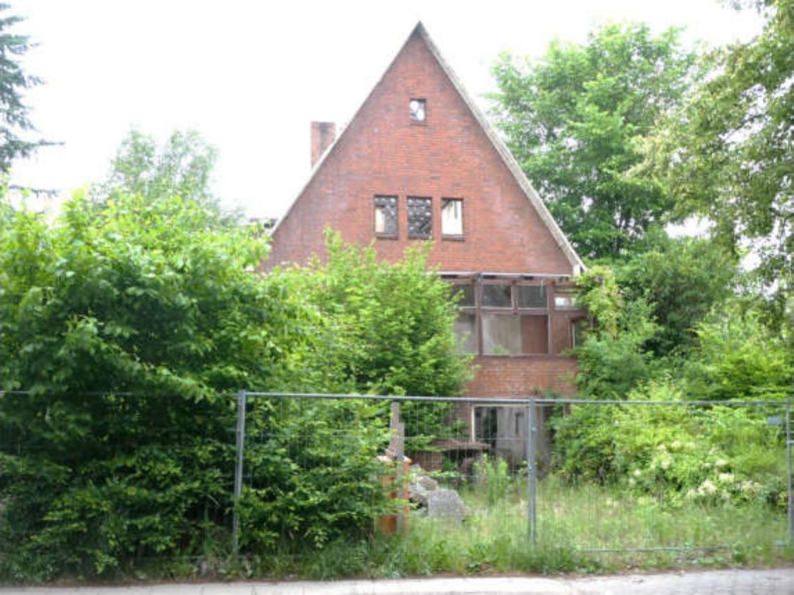 Vorderansicht eines zweistöckigen Backsteinhauses mit Satteldach, umgeben von üppiger Vegetation und einem Metallzaun