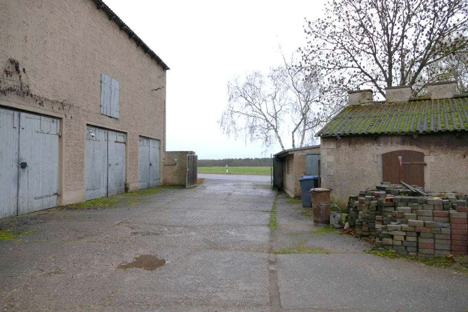 Gehweg zwischen zwei Gebäuden mit Garagen und Toren, rechts Stapel von Ziegeln, im Hintergrund Feld und kahle Bäume, Bebertal, Burgstraße, Auf der hohen Haide