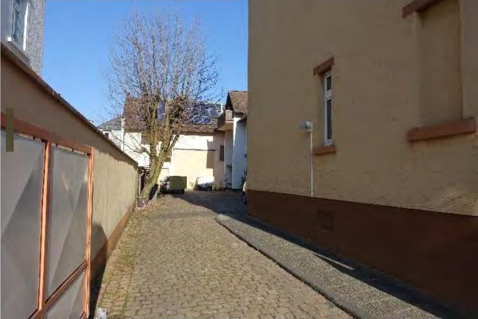 Narrower gepflasterter Durchgang zwischen zwei Gebäuden mit sichtbaren Außenwänden und Fenstern, im Hintergrund ein kahler Baum und weitere Gebäude