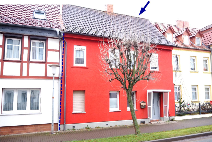 Vorderansicht eines roten Einfamilienhauses mit Satteldach, Fenstern und Eingang, umgeben von Nachbarhäusern und einem Baum vorne