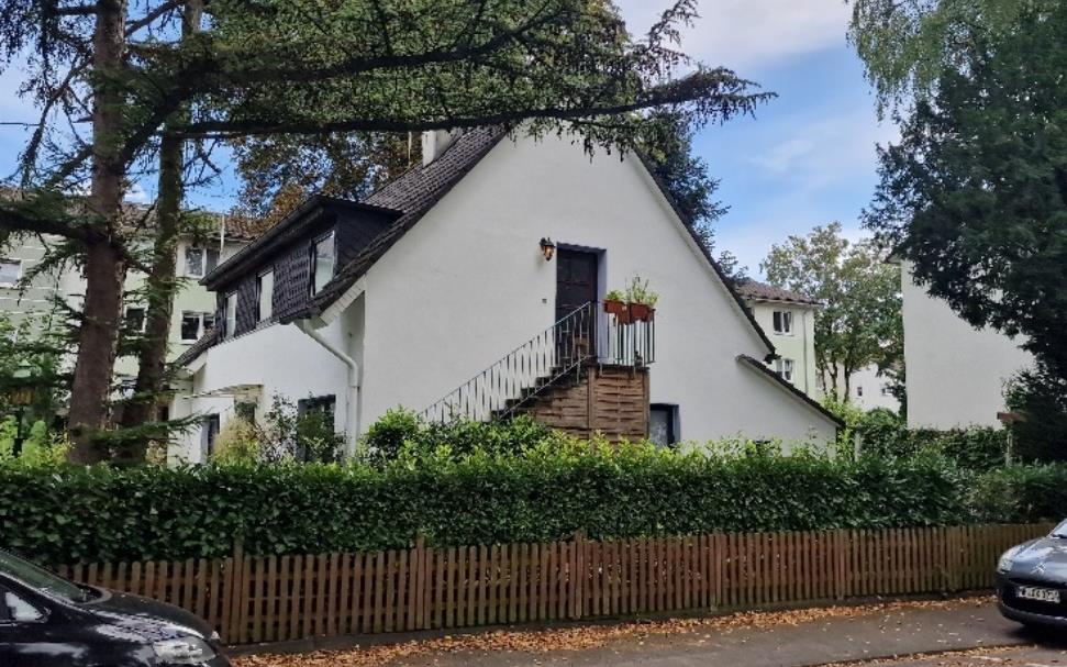 Vorderansicht eines Einfamilienhauses mit weißer Fassade, Eingangstreppe und Hecke, umgeben von Bäumen und geparkten Autos