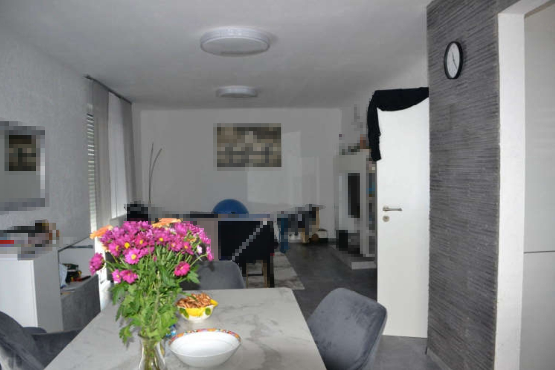 Innenraum eines Wohnbereichs mit Esstisch, Stühlen, Blumen und Blick in den Wohnraum mit TV-Regal und Wandbild