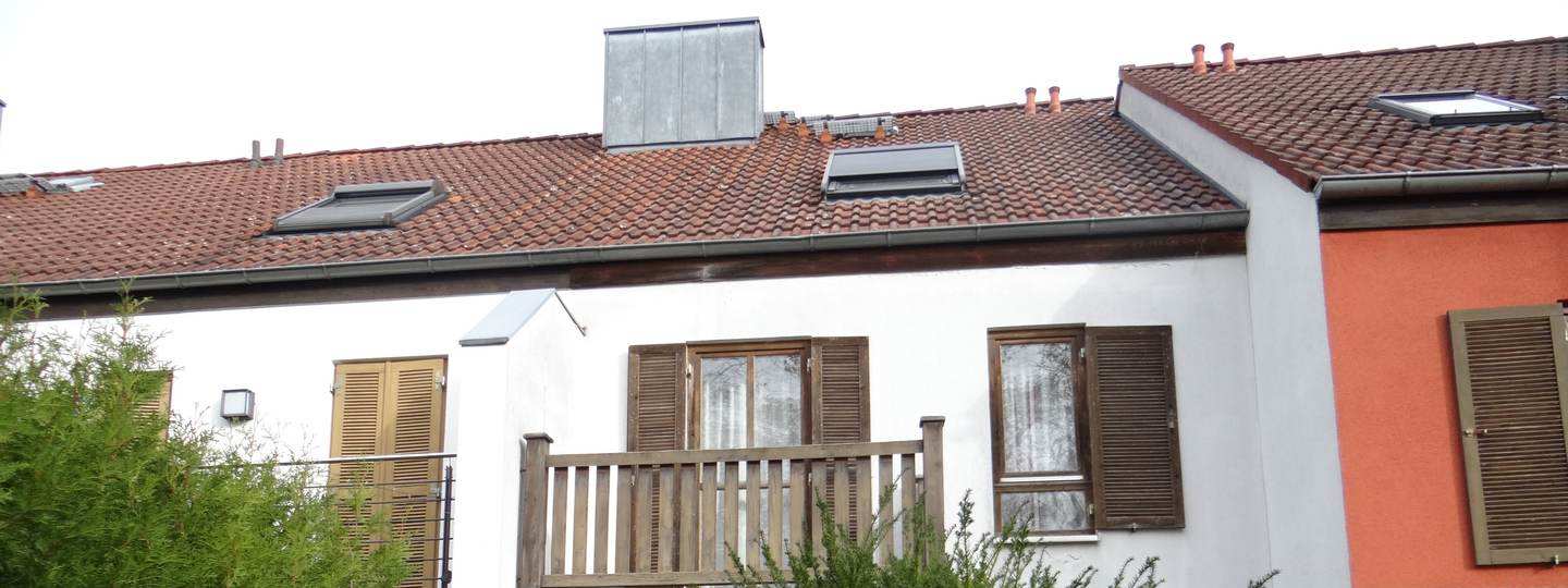 Rückansicht eines Einfamilienhauses mit rotem Ziegeldach, Fenstern mit Holzläden und einem Holzbalkon, umgeben von Grünanlagen
