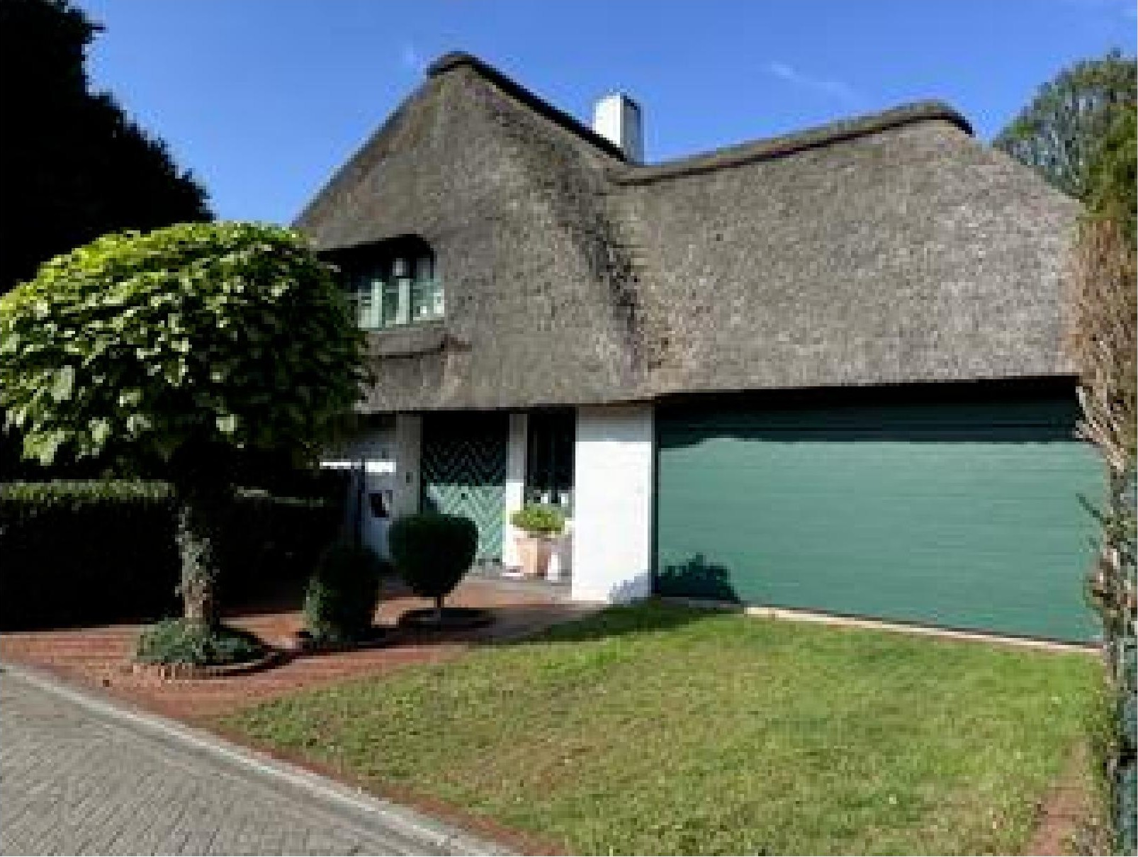 Einfamilienhaus mit Strohdach, Eingang, grüner Garagentür und Vorgarten mit gestutzten Bäumen und Rasen